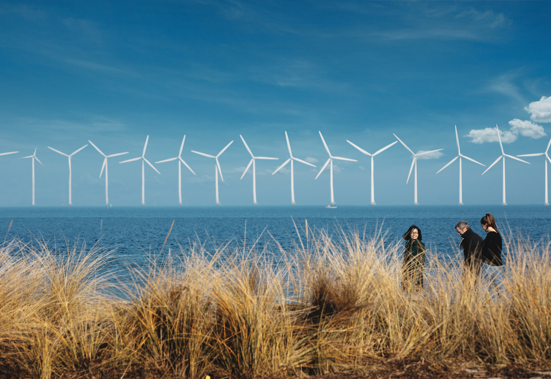 Three people walking on a beach, sea wind turbines in the background